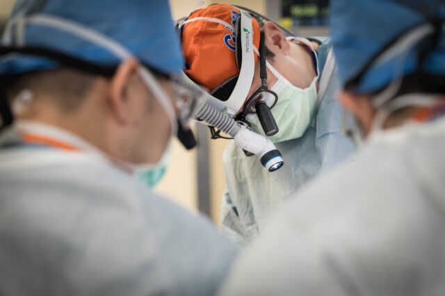 A group of surgeons perform an operation. The focus is on a male surgeon wearing a pair of glasses with surgical magnification lenses, a spotlight, and a head covering the Florida Gators logo.