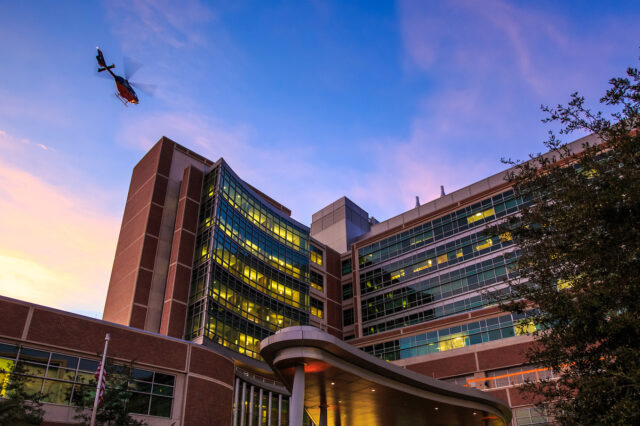 An orange and blue Shandscair helicopter gets ready to land on top of UF Health Shands Cancer Hospital. The sun is setting, and the building is surrounded by a blue sky and orange and pink clouds that resemble the University of Florida colors.