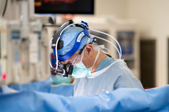 A doctor performs a surgery. He is wearing scrubs, a head covering, and a protective gown and is wearing glasses with magnifying glasses that allow him to see the surgical field more clearly. The patient is obscured by a sheet.