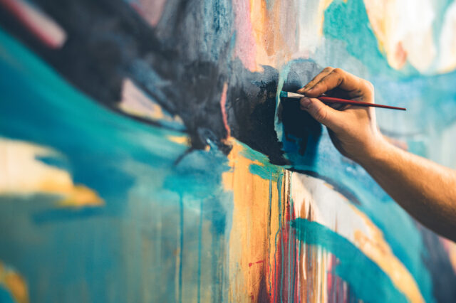 A hand is visible, hold a paintbrush.  The person is painting a colorful abstract canvas of blues, blue-greens, yellows and blacks.