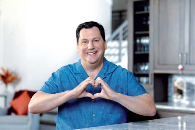 A man sits at a kitchen counter in his home. He is making the shape of a heart with his hands.
