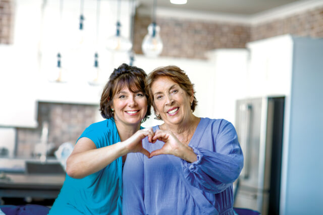 An older and younger woman stand side by side in a kitchen. They are forming the shape of a heart with their hands and they rest cheek to cheek.