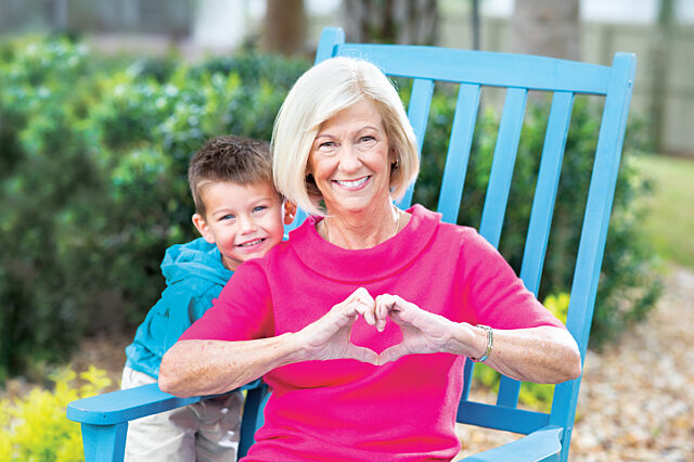 A woman sits in a blue chair in a garden. A little boy stands next to the chair and is leaning on her shoulder. The woman is making a stylized heart shape with her hands.
