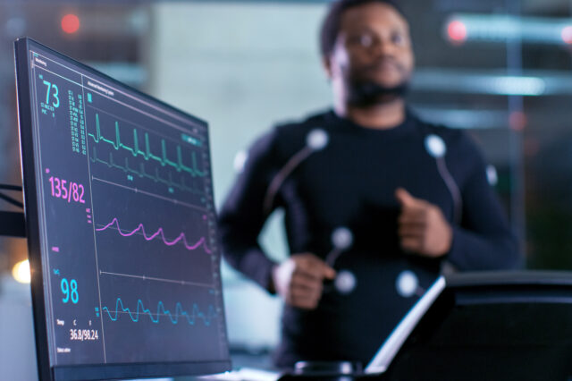 A man runs on a treadmill. He is wearing sensors on his chest, arms, and abdomen. A screen can be seen that is showing his vital signs as he runs.