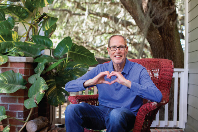 A man sits on a porch surrounding by green plants and trees. He is making the shape of a heart with his hands.