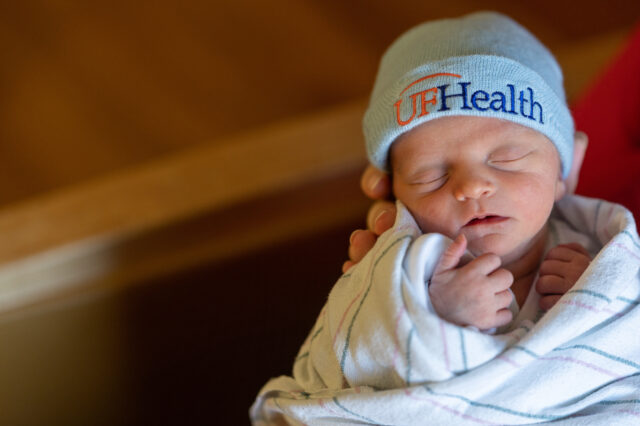 A newborn baby swaddled in a blanket and wearing a beanie that says UF health.