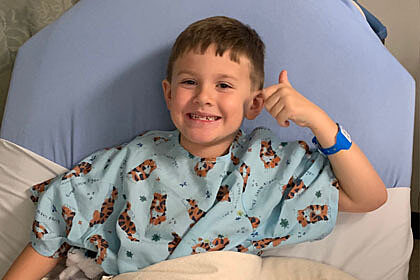 Baylor Bennett wearing a hospital gown tucked into a hospital bed smiling and giving a thumbs up