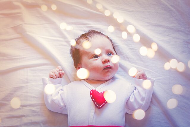 A baby looking up at lights, wearing a jumper with a felt heart on it.