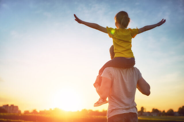 A child sits on a man's shoulders, looking at the setting sun, They are holding the arms out wide.
