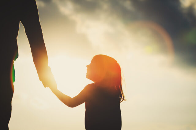 A girl holds and adult's hand, silhouetted by a sun in a cloudy sky.