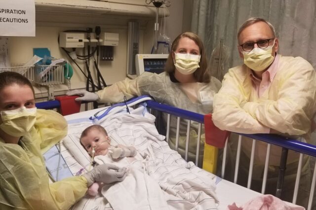 A small child lays in a hospital bed. She is surrounded by adults wearing yellow protective gowns and yellow face masks.