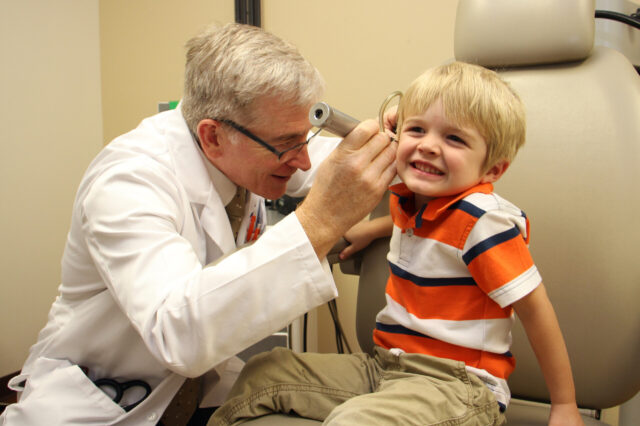 A photograph of a friendly, gray haired male doctor checking the right ear of a smiling, blond haired boy who is about five years old. Represents a new UF Health study questions the safety of antibiotic ear drops prescribed to children after ear tube surgery.