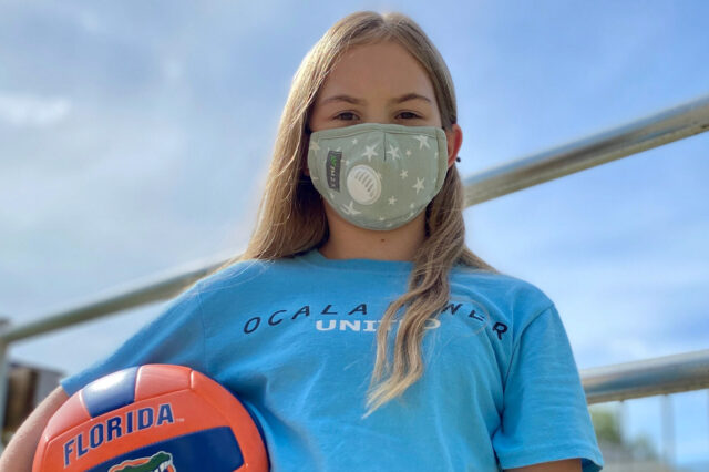 Kendall Lewis stands next to a volleyball net on a sunny day. She is wearing a face mask and is carrying an orange and blue Florida Gators volleyball.