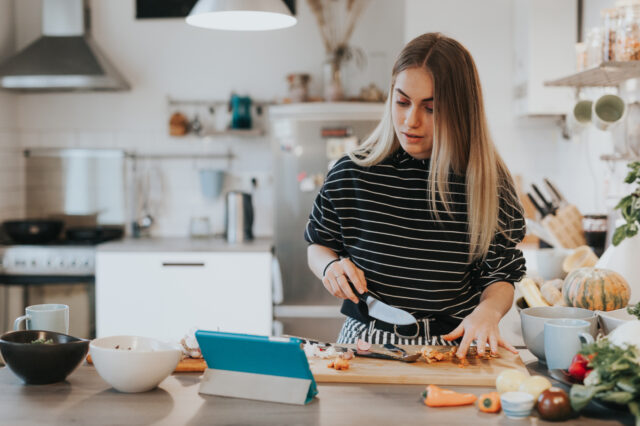 A woman looks at her laptop while making a meal in her kitchen.