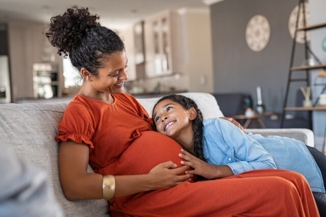 A child lays on her mother's pregnant belly as they smile at one another.