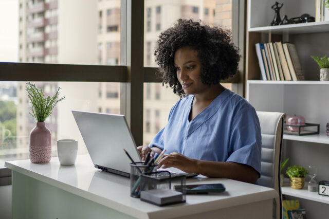 A woman wearing blue scrubs sits at a desk in an office. She is sing a computer while taking notes.