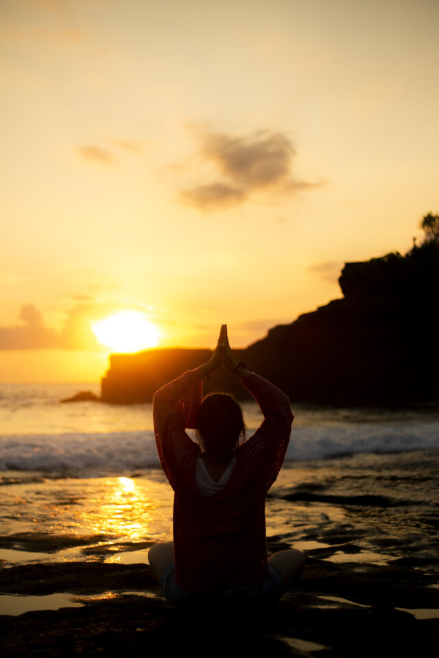 A woman practices yoga on a beach in the light of the setting sun.