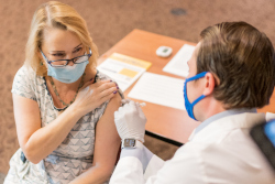 A woman sitting down wearing a mask rolls up her sleeve while a man, also wearing a mask, giver her a vaccine shot. 