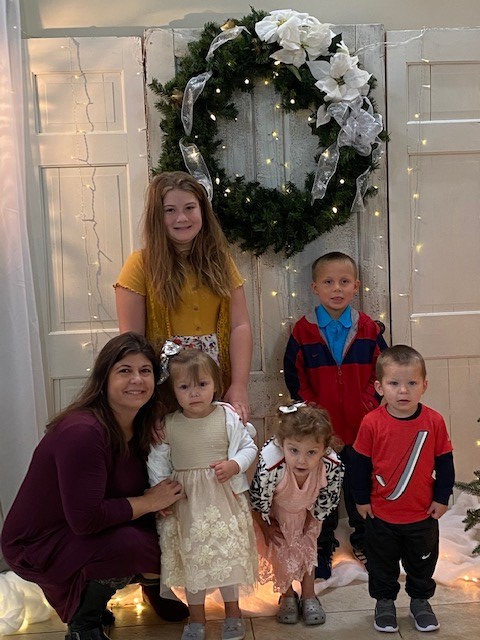Kim smiling at the camera with her daughter and grandchildren standing against a wooden door that features a Christmas wreath. 