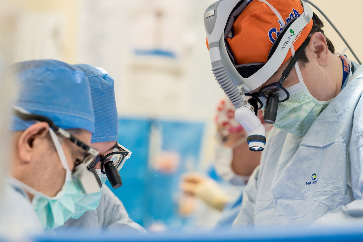 Medical professionals in blue scrubs perform a liver transplant in an operating room. Dr. Thiago Beduschi wears a Florida Gators surgical cap and is wearing a pair of specialized glasses that magnify the surgical area.