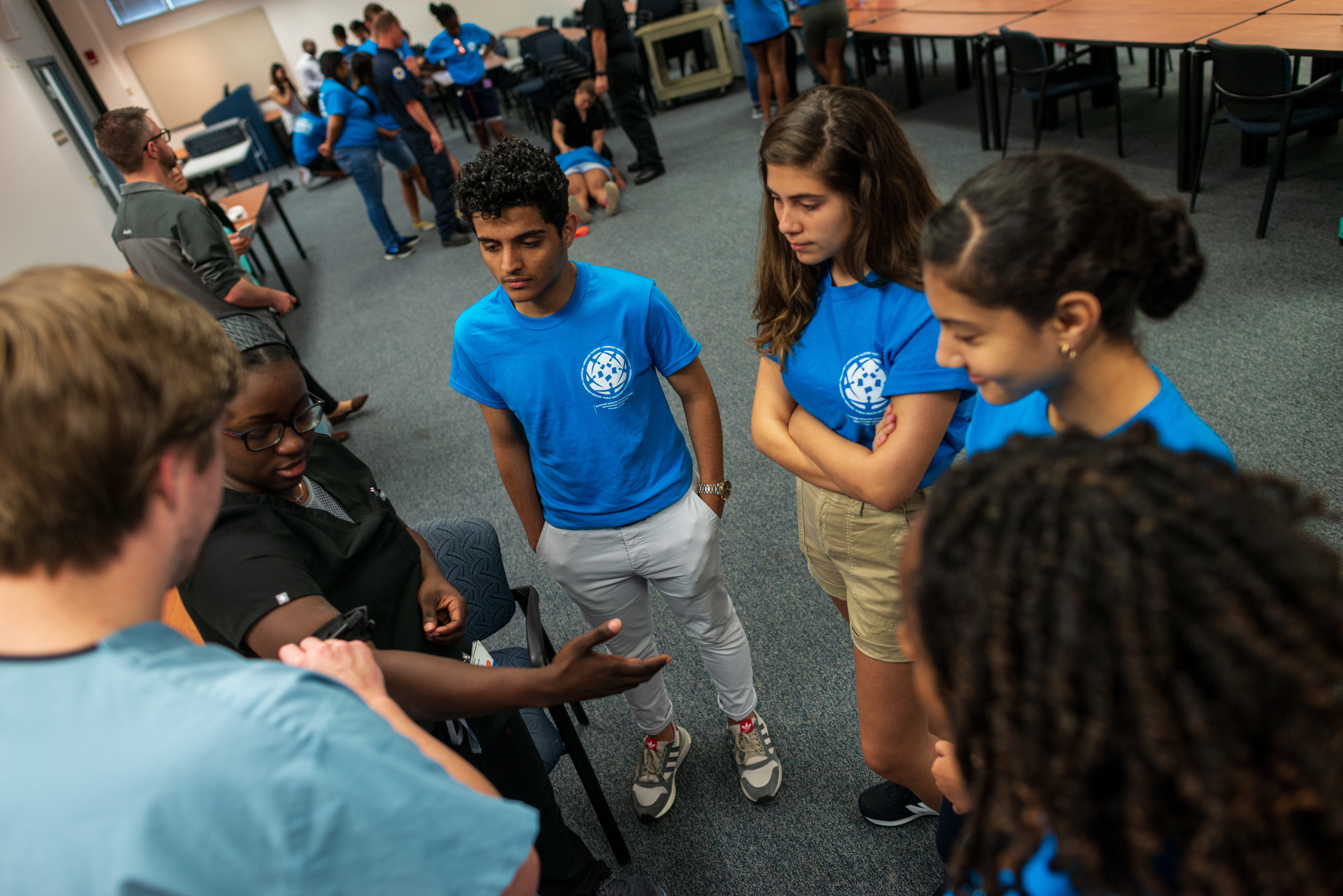 Students in matching tshirts gather around a person in scrubs demonstrating a medical procedure on another person in scrubs