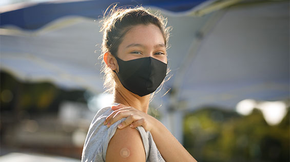 A teenage raises her shirt sleeve to show us her vaccination bandage. She is wearing a face mask.