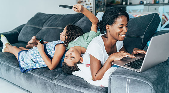 A young Black mother works on a laptop on her couch. Two children lay across her back, looking at their phones. 