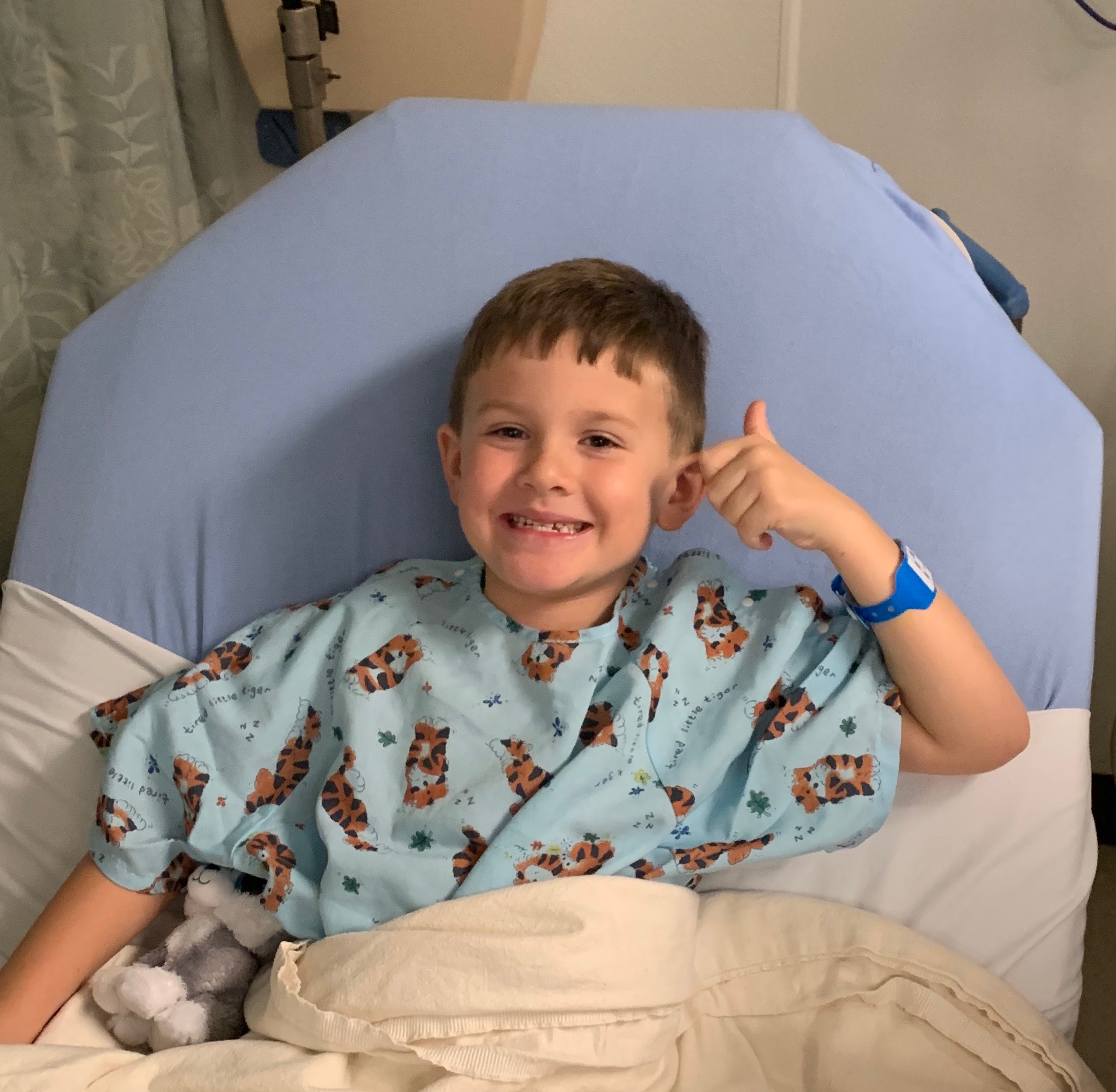 baylor in a hospital gown tucked into a hospital bed while smiling and holding a thumbs up