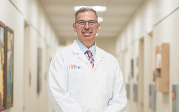 Dr. Michael Jaffee stands in the middle of a clinic hallway. He is wearing a white UF Health medical coat and a plaid tie with the orange and blue colors of the University of Florida.