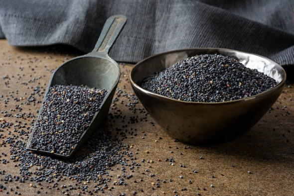A bowl of dark, black poppy seeds sits on a wooden surface. Next to it is a scoop also full of poppy seeds.