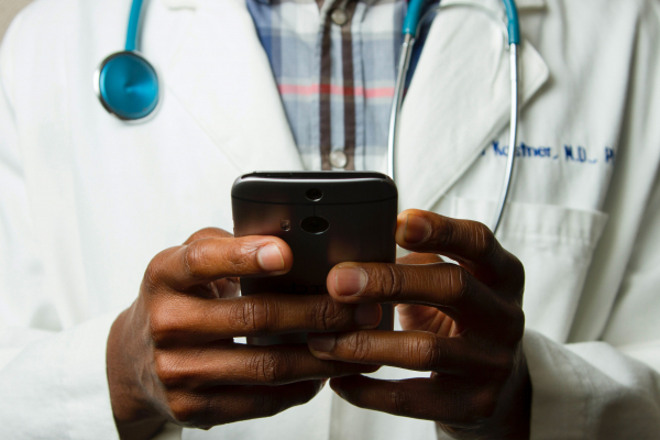 A male Black physician wearing a white medical coat looks at his smartphone.