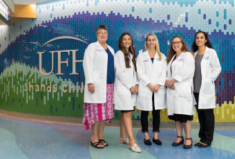 Five midwives, wearing white UF Health medical coats, stand in front of the tiled lobby wall in the UF Health Shands Children's Hospital.