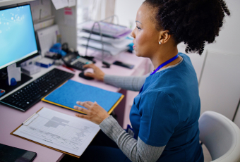 A woman wearing blue medical scrubs is sitting at a computer, inputting information from notes.