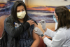 Emily Benitez lifts the sleeve of her outfit to receive an injection of the Moderna COVID-19 vaccine. A woman wearing a white medical coat provides the injection. Both are wearing masks.